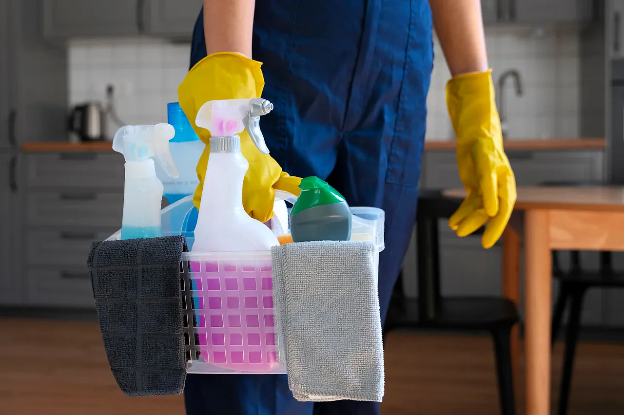 Profissional de luvas amarelas carregando cesto com produtos de limpeza